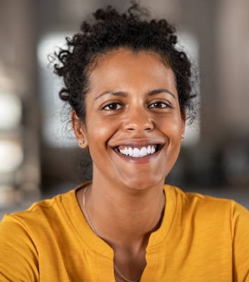 Smiling woman with curly hair wearing a yellow shirt, sitting in a bright indoor space.