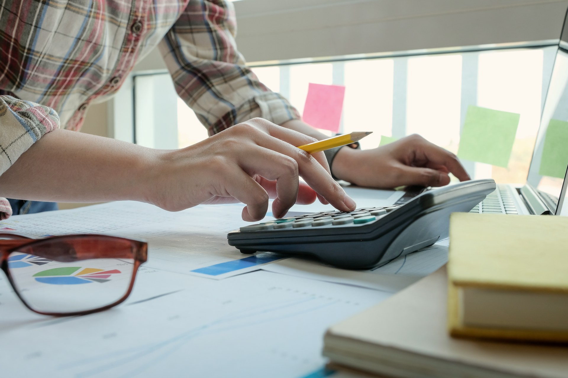 Hands using a calculator with notes, glasses, and a laptop on a work desk.